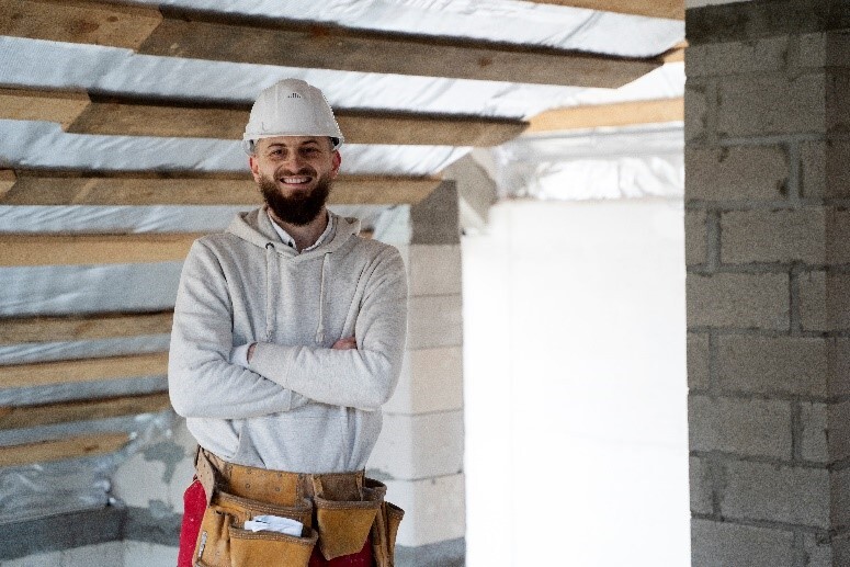 Comprehensive Guide to Roofing Services in Atlanta 11 Smiling bearded man in a white hard hat and grey hoodie with arms crossed, wearing a tool belt, standing inside a construction site.
