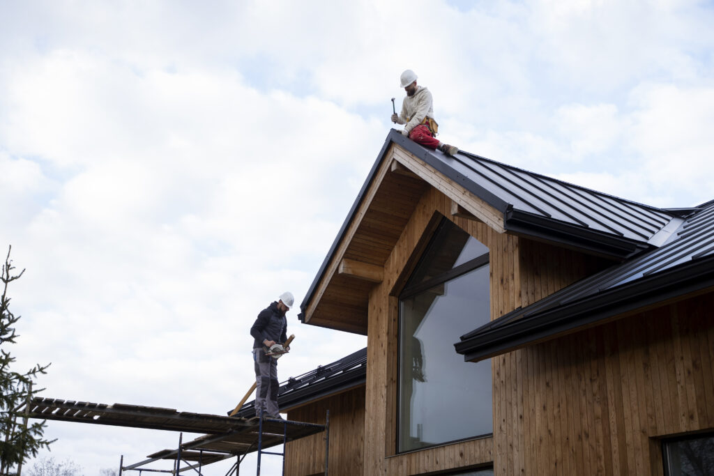 Complete Guide to Residential Roofing Services in Atlanta 3 A roofer with a beard, wearing a white helmet and earbuds, examines a nail while sitting by a dark metal roof and wooden facade.