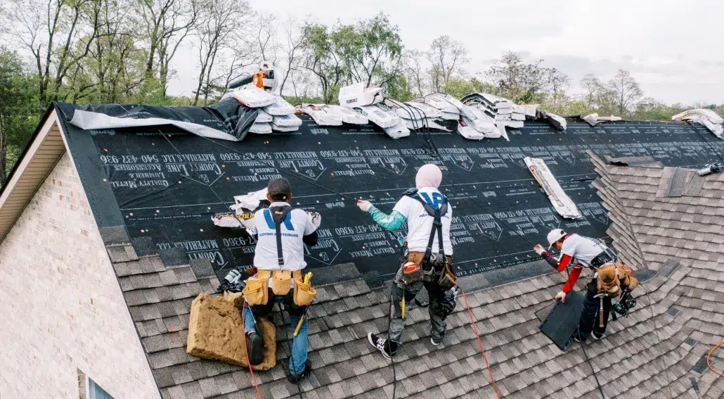 Roofing crew installing new shingles on a steep roof, with underlayment partially covered and stacks of shingles near the ridge.