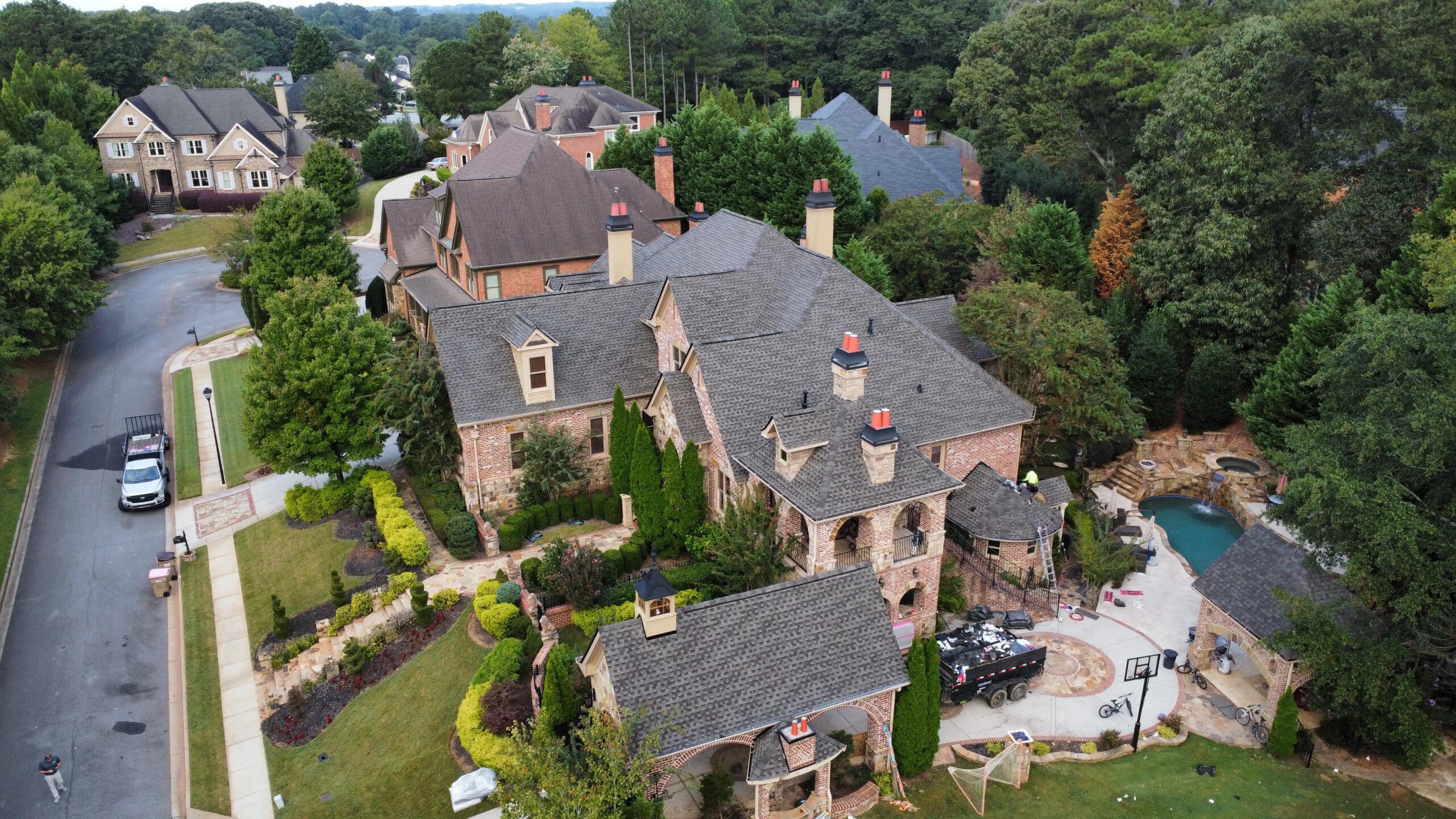 Aerial view of a large brick luxury home with multiple gables and chimneys, landscaped yard, and backyard pool area, located in an upscale suburban neighborhood.