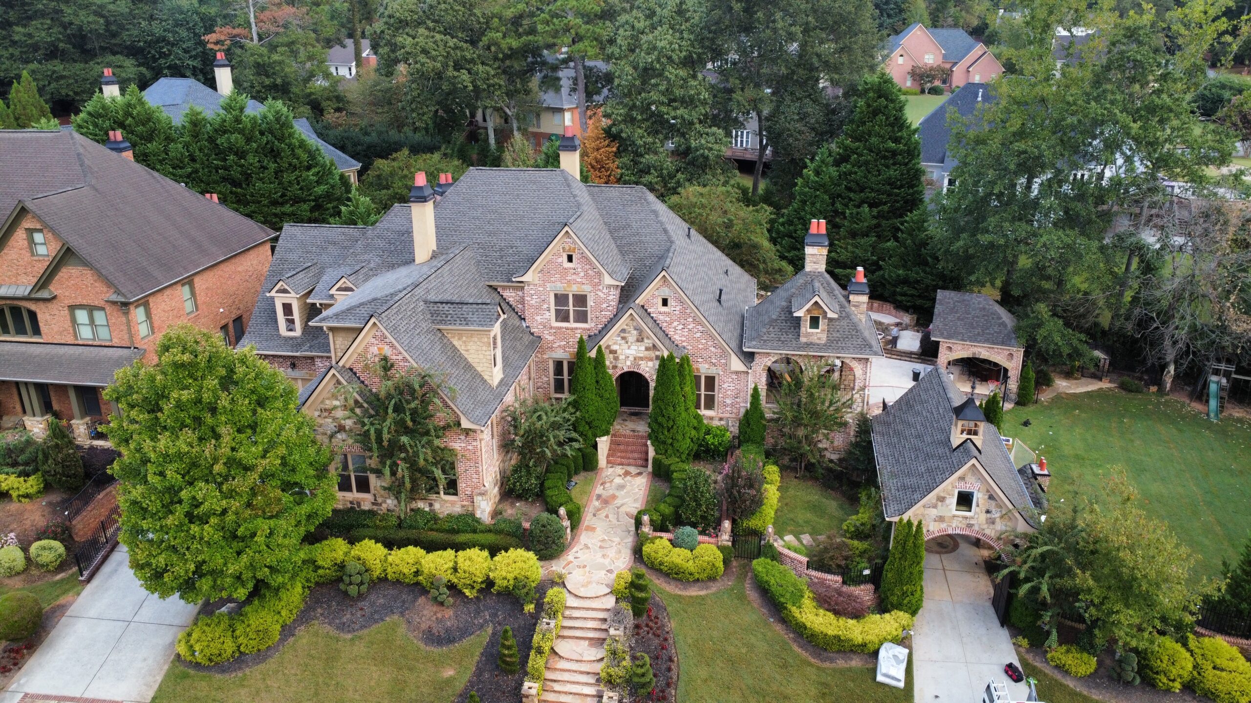 Aerial view of a large brick and stone luxury home with multiple gables and chimneys, surrounded by manicured landscaping, trees, and neighboring houses in a suburban neighborhood.