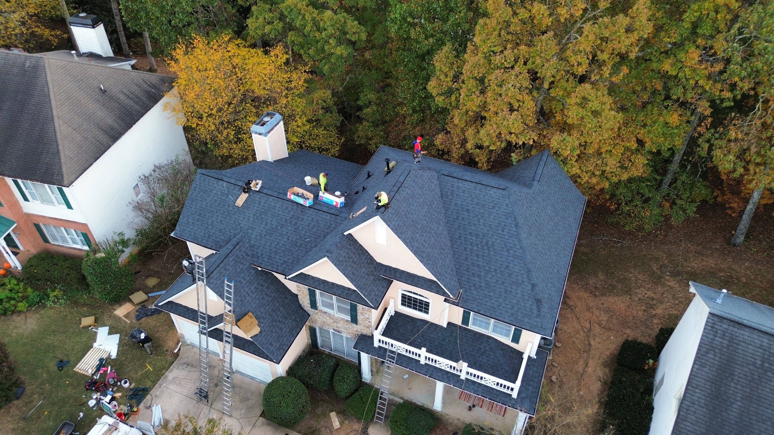 Aerial view of workers installing a new dark gray shingle roof on a two-story suburban house surrounded by trees with autumn foliage.