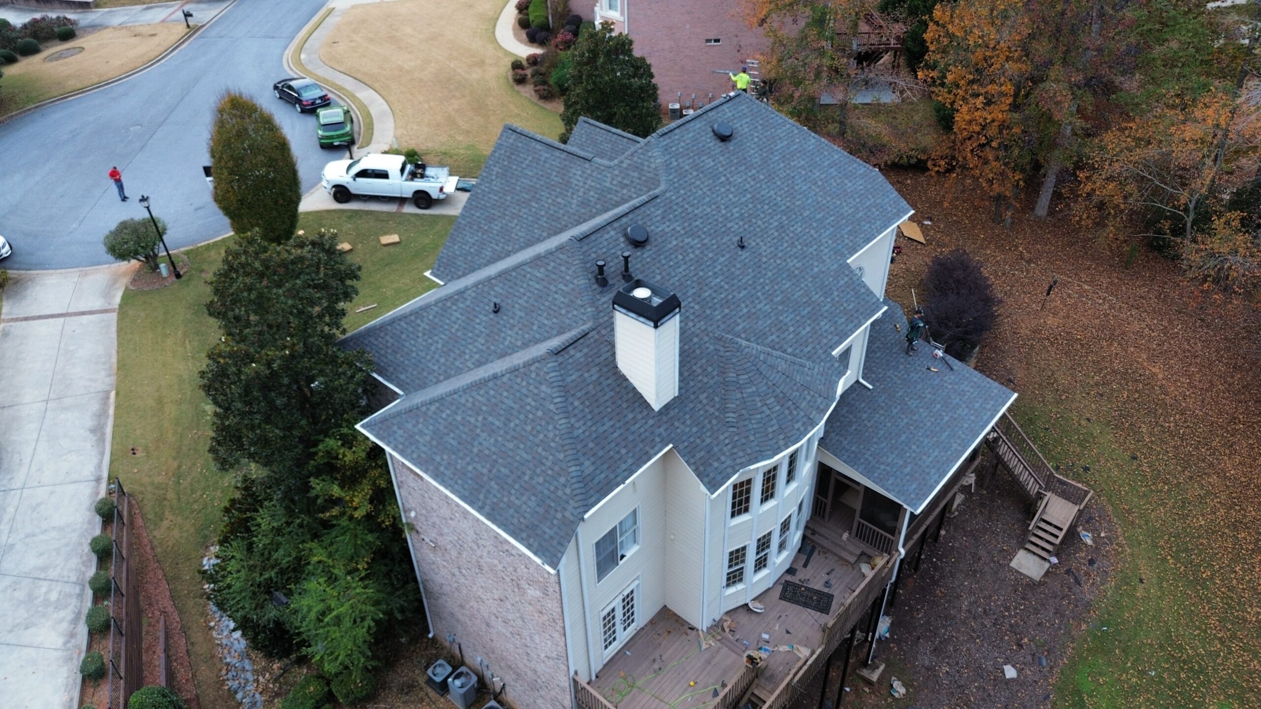 Aerial view of a large two-story suburban house with a new gray shingle roof, surrounded by trees and lawns, with vehicles and workers visible nearby.