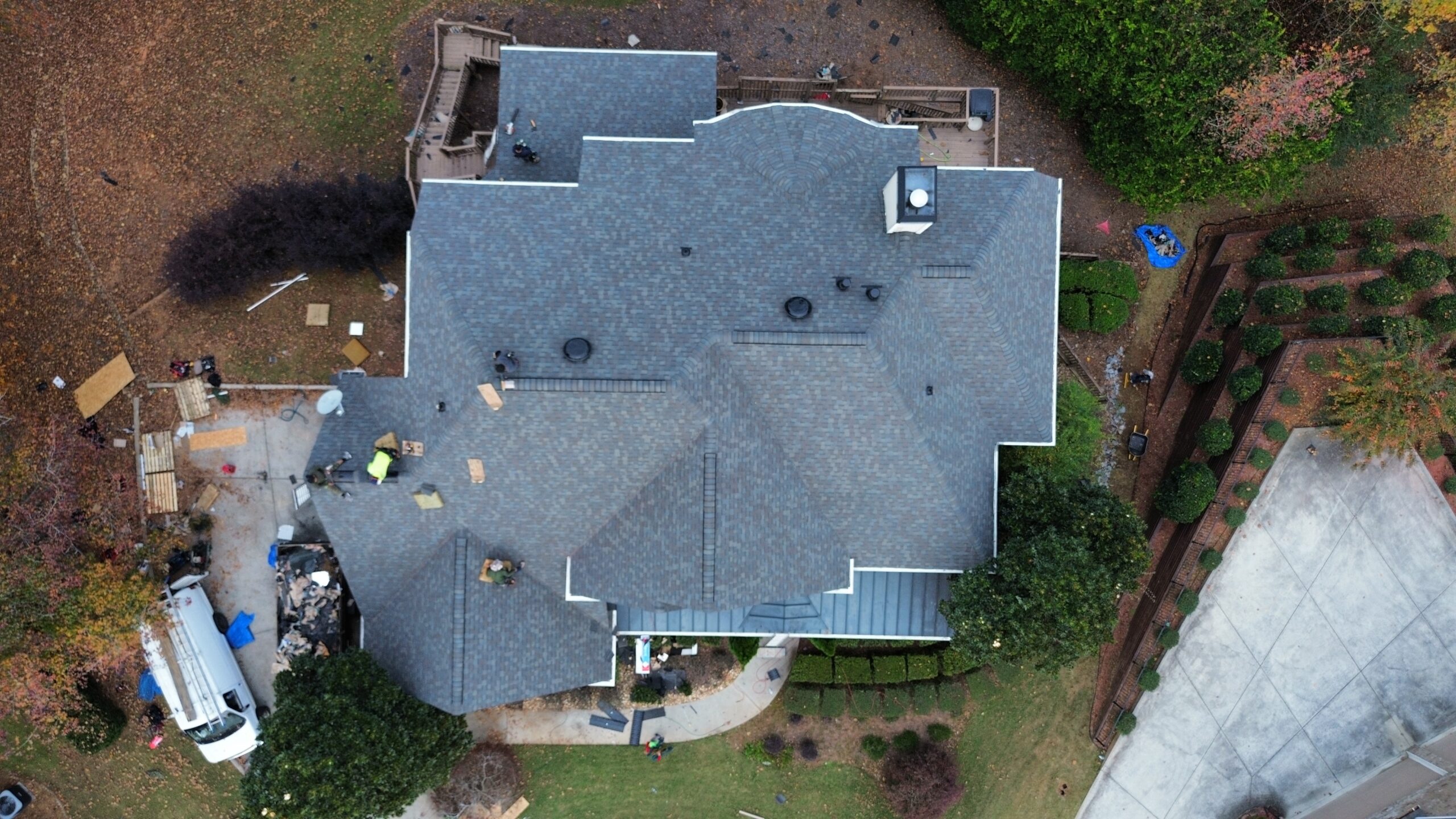 Aerial top-down view of a house with a new gray shingle roof, showing workers, equipment, and vehicles around the property during roof installation.