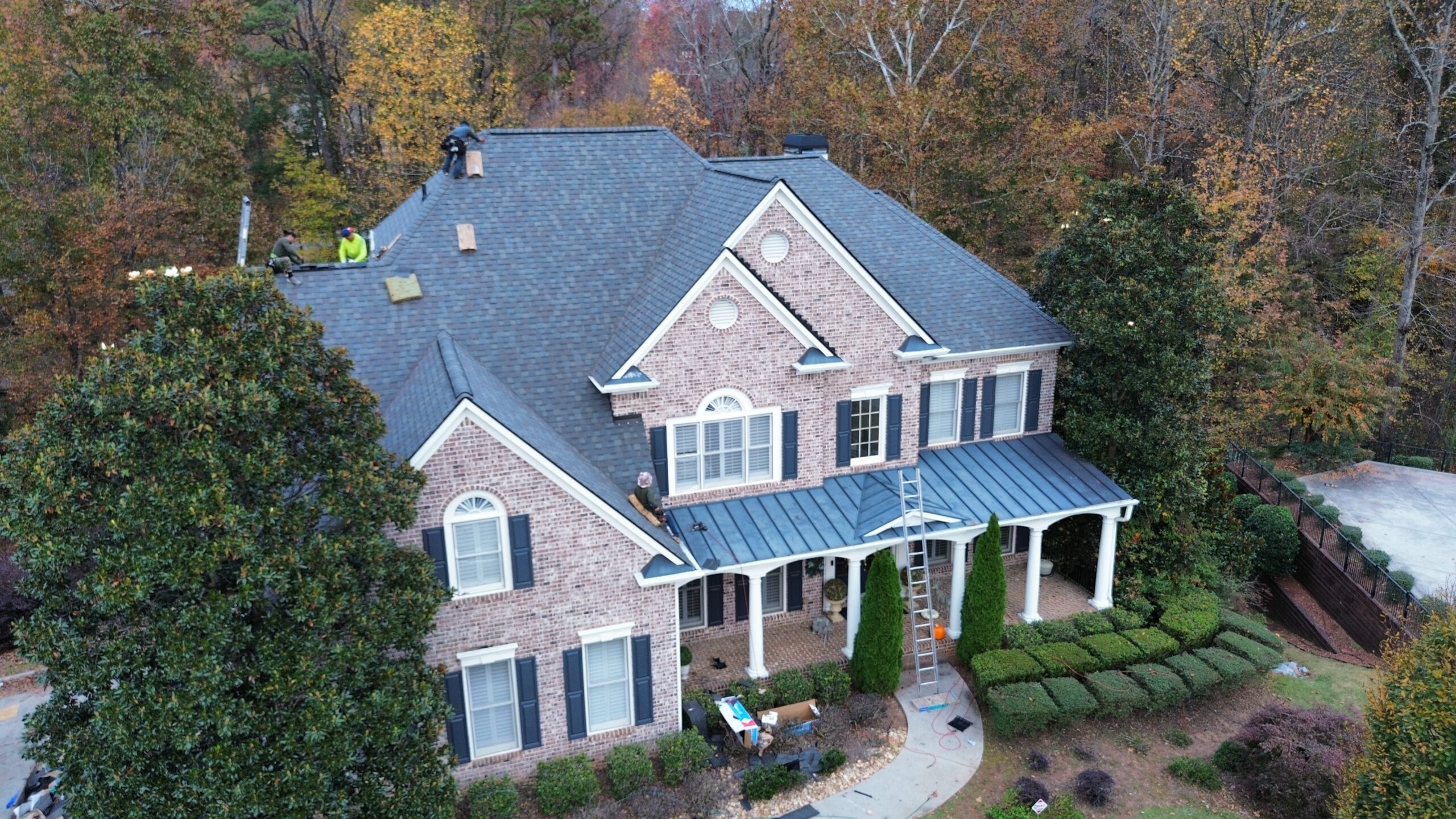 Aerial view of a two-story brick house with workers installing a new gray shingle roof, surrounded by trees with autumn foliage.