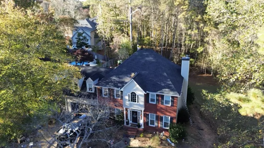 Aerial view of a two-story brick house with a new dark gray shingle roof, surrounded by trees and a wooded backyard.