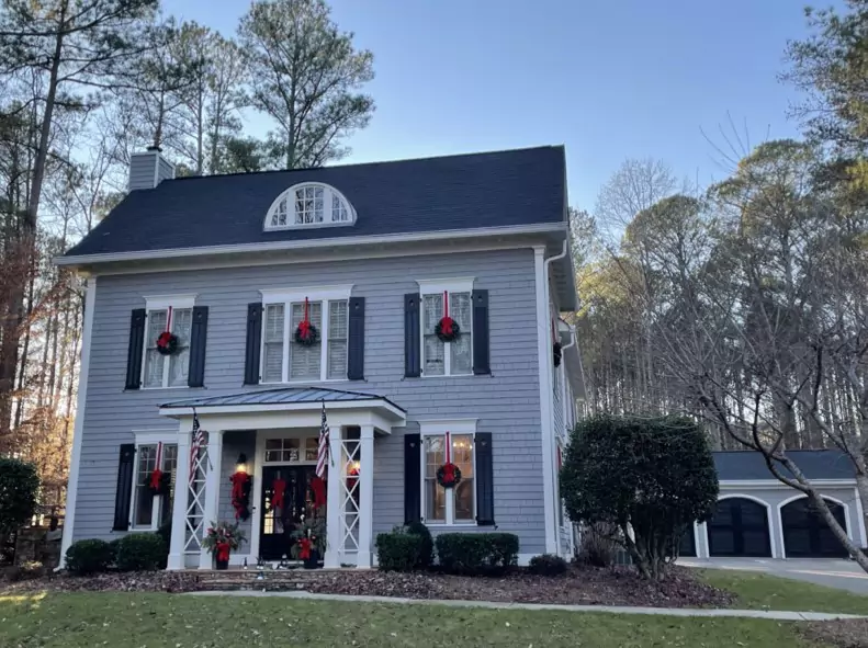 Two-story gray house with black shutters and a black roof, decorated with red wreaths and ribbons for the holidays, surrounded by trees and a detached garage.